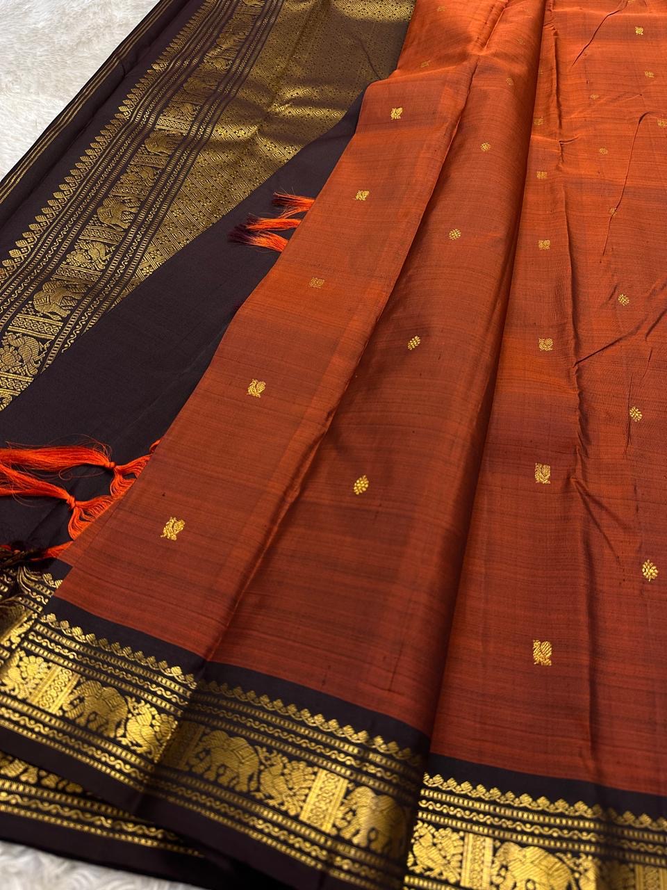 Angled close-up of rust Pure Kanchipuram silk saree with small gold zari buttas, black border, and orange tassels beside the pallu.