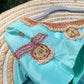 Turquoise fabric with intricate gold and pink embroidery on a hanger against a blurred green background.