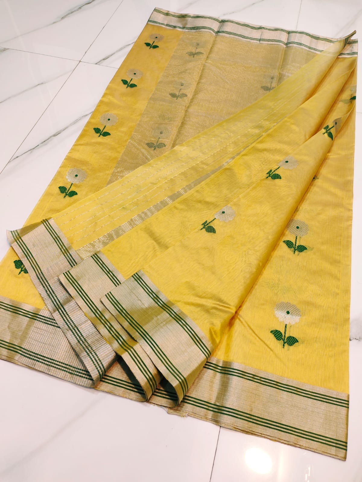 Folded Turmeric Yellow Pattu Silk Chanderi saree showing silver floral motif with green woven leaves and metallic striped border